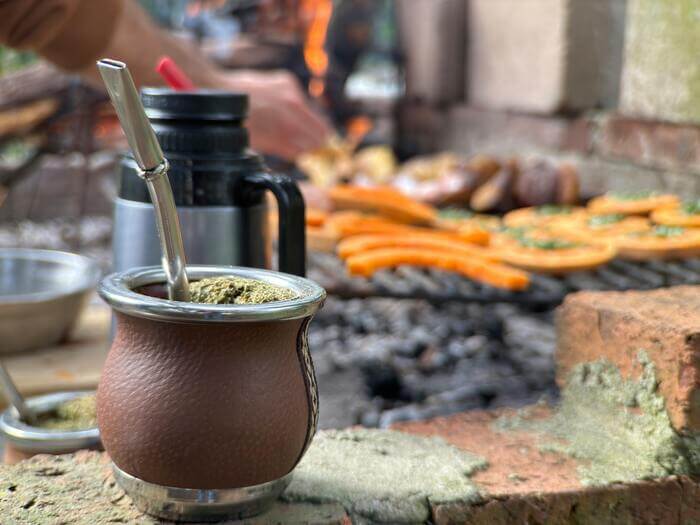 Cuia de chimarrão em primeiro plano com churrasco assando ao fundo, representando a tradição do Gaúcho